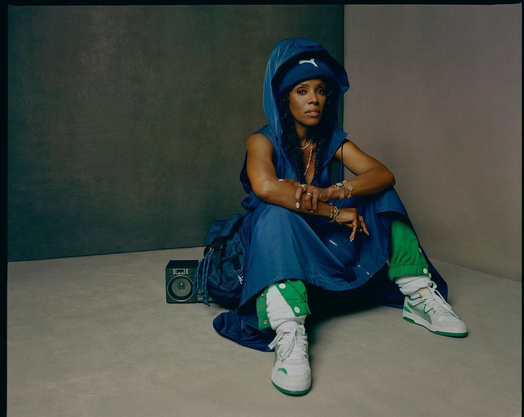 African American woman in blue hooded dress sitting on floor with boombox and backpack, wearing white and green sneakers in studio.