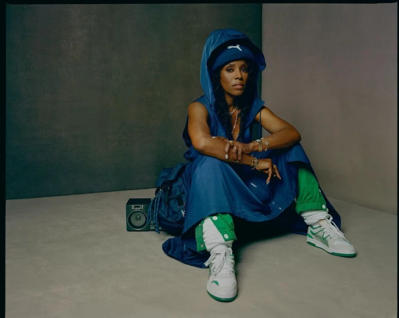 African American woman in blue hooded dress sitting on floor with boombox and backpack, wearing white and green sneakers in studio.