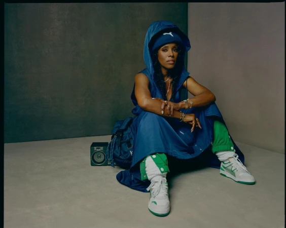 African American woman in blue hooded dress sitting on floor with boombox and backpack, wearing white and green sneakers in studio.