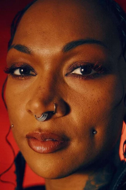 Close-up portrait of an adult woman with a nose ring, visible tattoo, and red background. Beauty shot with warm color grading.