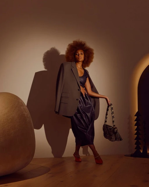 Fashionable woman with an afro poses in a studio wearing a blazer and a dress, holding a patterned handbag and wearing red shoes.
