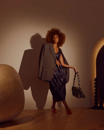 Fashionable woman with an afro poses in a studio wearing a blazer and a dress, holding a patterned handbag and wearing red shoes.