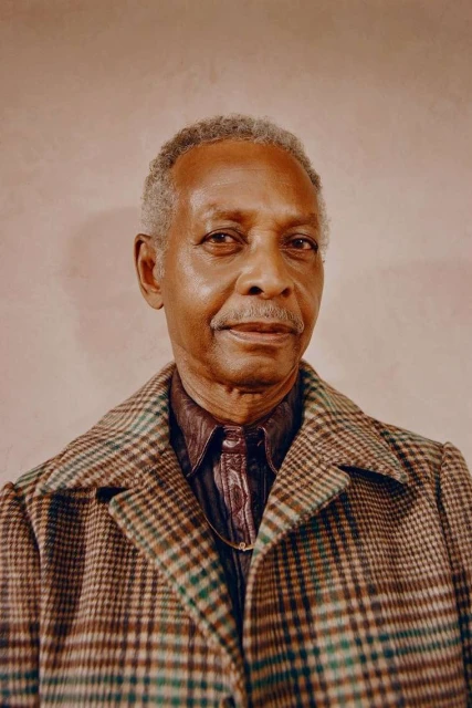 Portrait of an elderly African-American man with graying hair wearing a patterned brown coat and collared shirt against a peach backdrop.