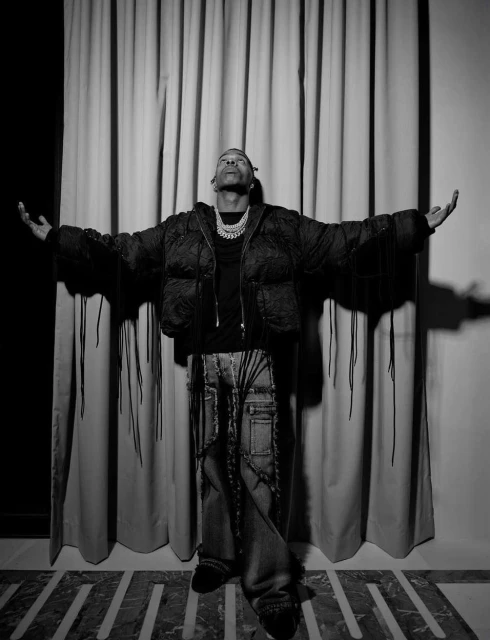 A single man with arms outstretched wears a jacket and jeans, posing in front of a curtain in a black and white studio portrait.