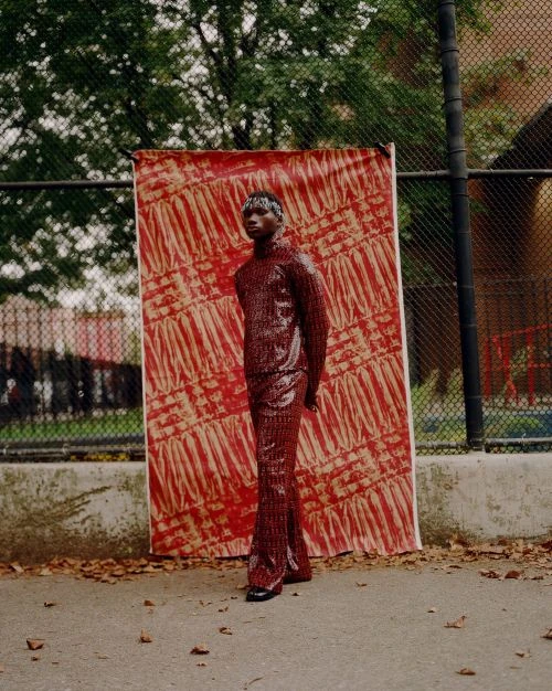 Dark-skinned adult in a red sequin turtleneck and flared pants, standing in front of a patterned backdrop on an outdoor basketball court.