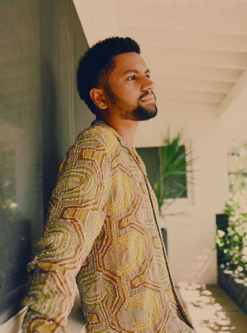 Man in patterned jacket poses against a wall with natural light streaming in, showcasing a blend of fashion and architectural detail.