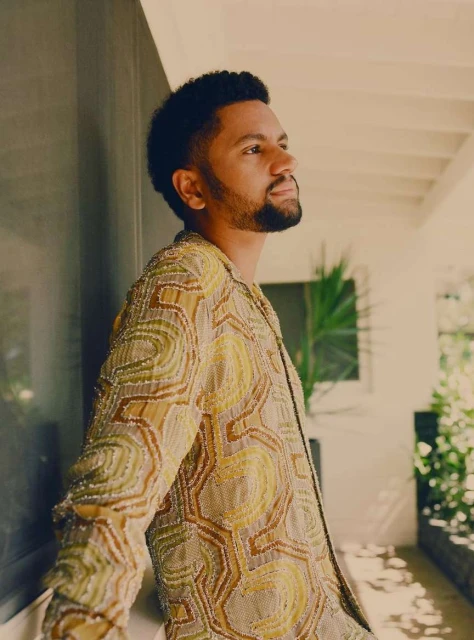 Man in patterned jacket poses against a wall with natural light streaming in, showcasing a blend of fashion and architectural detail.