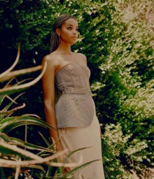 A woman wearing a strapless metallic top and skirt standing outdoors amidst lush green foliage with sunlight filtering through leaves.