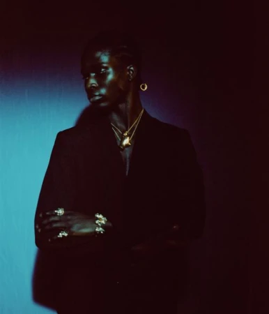Portrait of a black man with braided hair wearing a dark blazer and gold jewelry against a dark blue and black background.