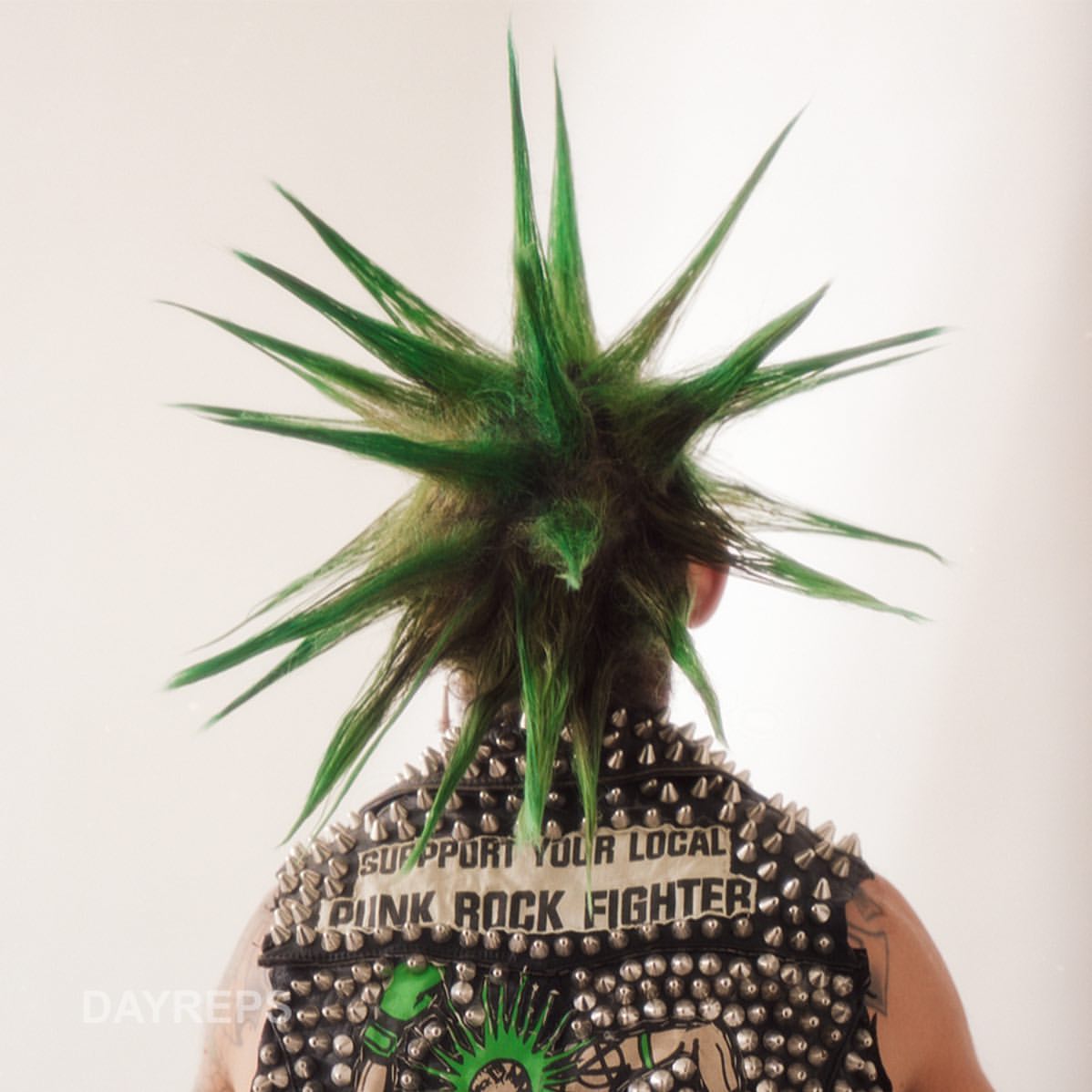 Person with green spikey hair and studded jacket facing away from the camera in a studio setting; punk fashion, vibrant colors, edgy style.