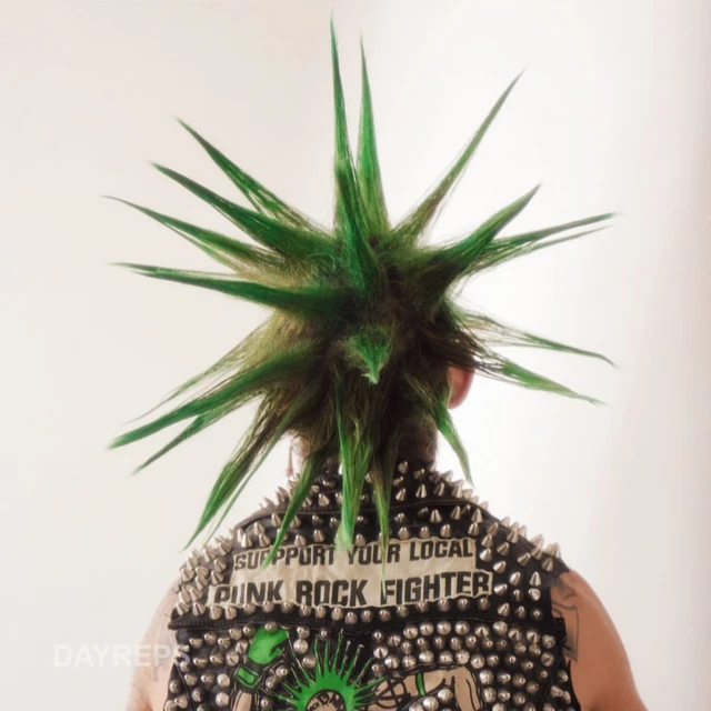 Person with green spikey hair and studded jacket facing away from the camera in a studio setting; punk fashion, vibrant colors, edgy style.