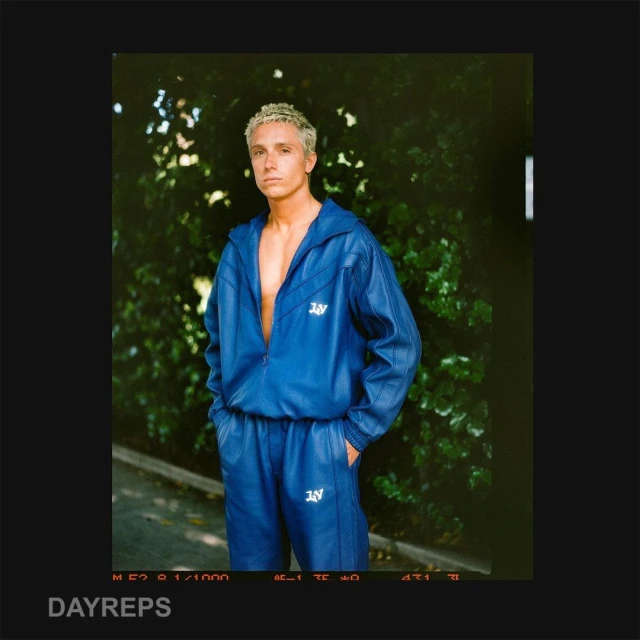 A fair-skinned adult male with blonde hair poses in a blue tracksuit against a green leafy backdrop, outdoors in natural lighting.