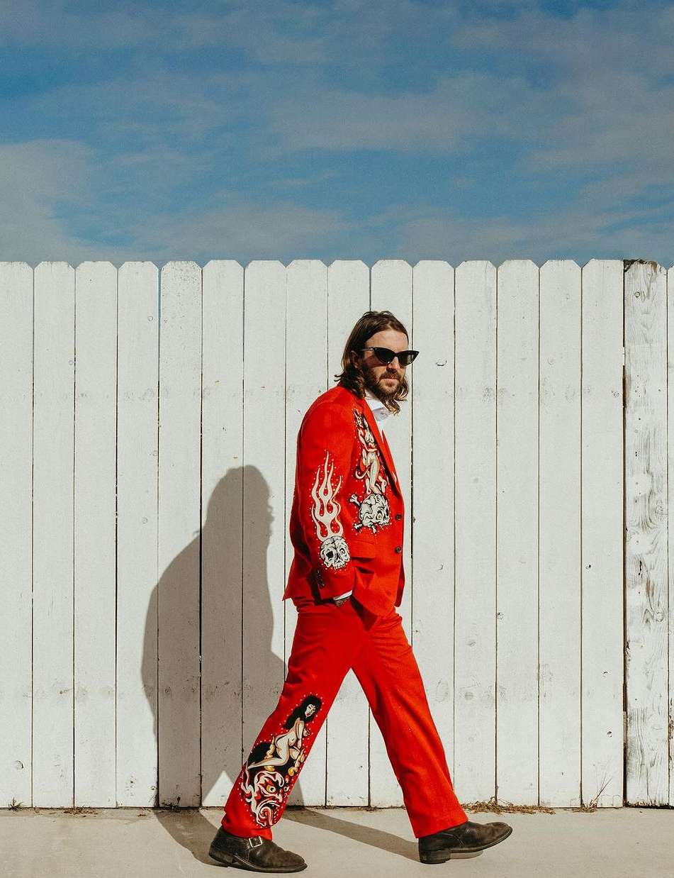 Man in red patterned suit walks past a white fence under a blue sky. The suit features skull, flame, and nude figure design details.