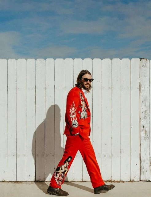 Man in red patterned suit walks past a white fence under a blue sky. The suit features skull, flame, and nude figure design details.
