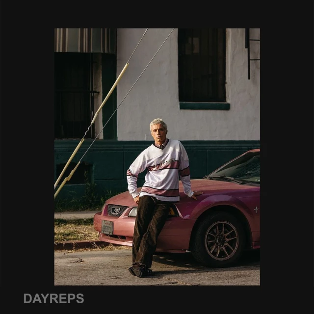 Adult male model leans against a pink Ford Mustang on a city street, posing in a white hockey jersey and dark jeans.