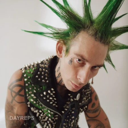 Portrait of a young adult male with a green mohawk, tattoos, and a studded vest, posing indoors against a neutral background.