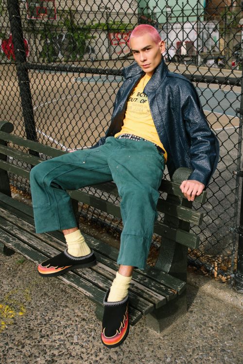 Young adult with pink hair is sitting on a park bench wearing a blue jacket, yellow t-shirt, and green pants with flame shoes.