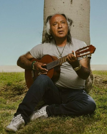 Adult male sitting on grass, playing an acoustic guitar next to a tree on a sunny day with a blue sky background.