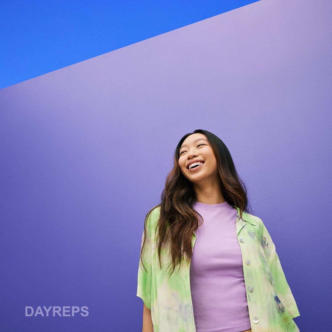 Smiling Asian woman in casual fashion stands against a purple wall with a blue accent, wearing a green tie-dye shirt, and purple top.