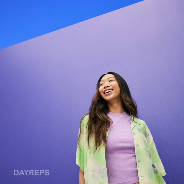 Smiling Asian woman in casual fashion stands against a purple wall with a blue accent, wearing a green tie-dye shirt, and purple top.