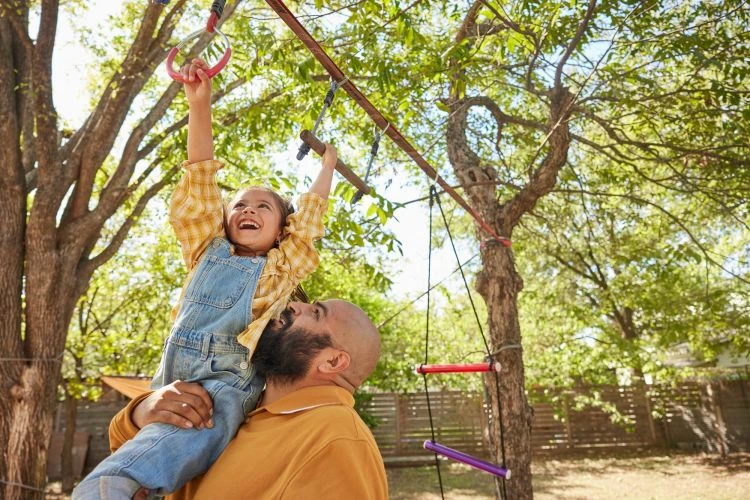 A smiling child in overalls is held up by an adult and reaches for a hanging ring on a backyard climbing structure under green trees.