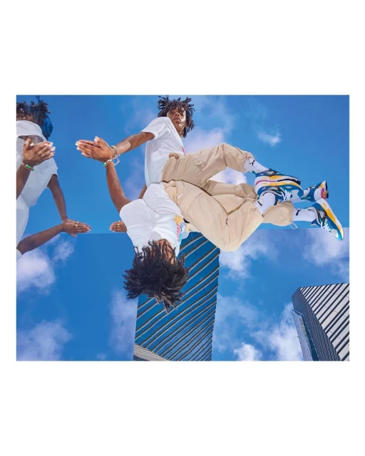 Person in white t-shirt and tan pants poses near mirror reflecting buildings and blue sky. The focal point is fashion sneakers.