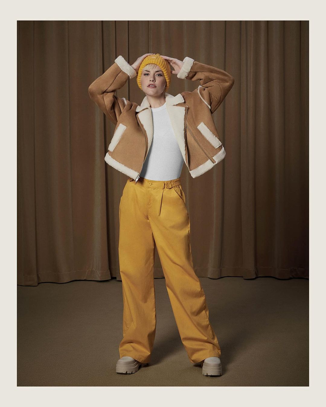 Full-length studio shot of a woman posing in a brown shearling jacket, yellow knit beanie, white top, and yellow pants against a brown curtain backdrop.