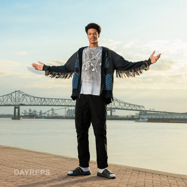 A young adult stands confidently in a stylish fringed jacket by the river with a bridge in the background under a clear, bright sky.
