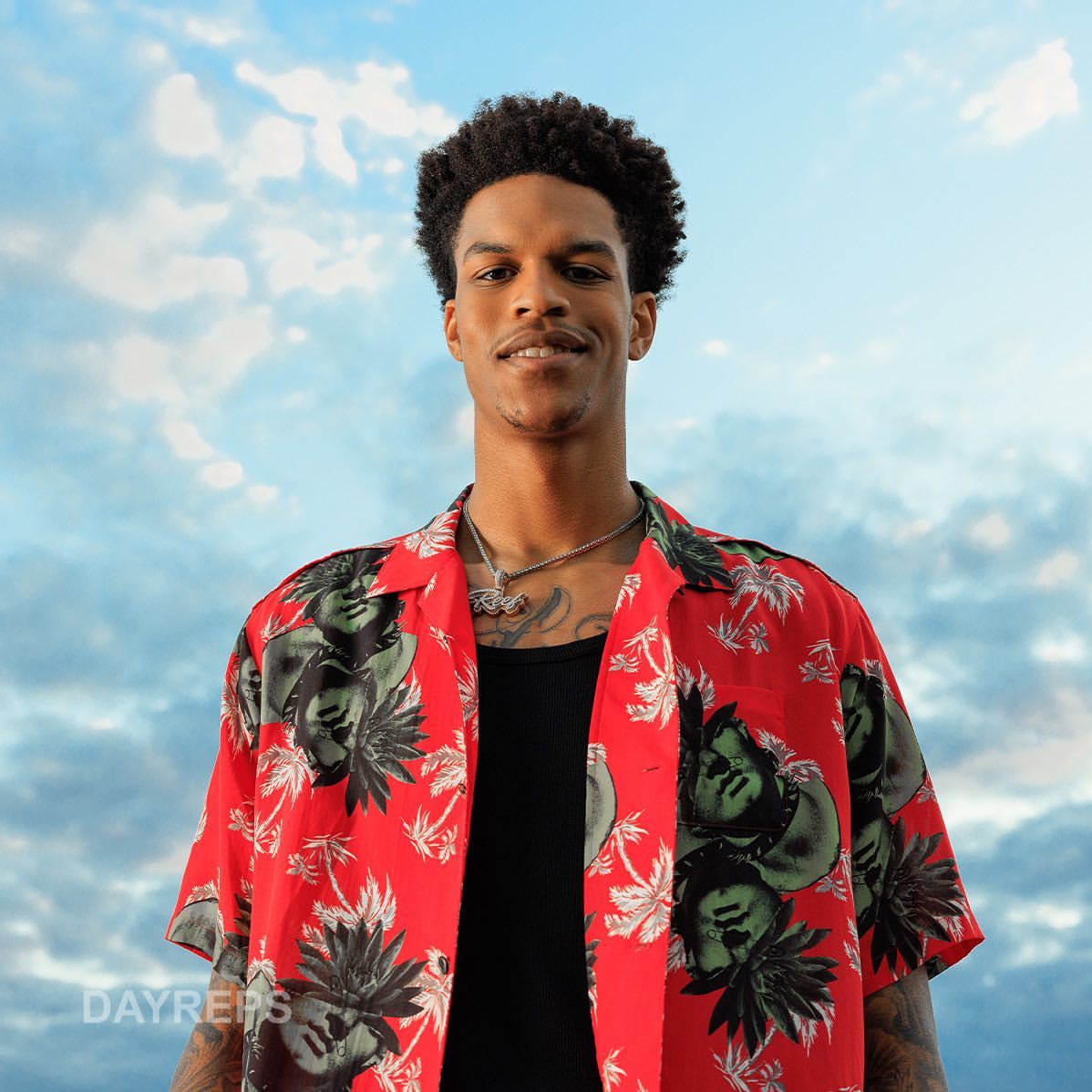 Portrait of a smiling black man wearing a red patterned short-sleeve shirt against a cloudy blue sky.
