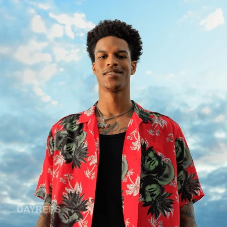 Portrait of a smiling black man wearing a red patterned short-sleeve shirt against a cloudy blue sky.