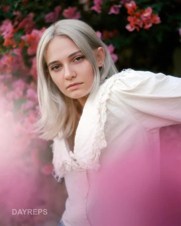 Portrait of a blonde woman wearing a white lace-collared blouse in front of pink flowering bushes by Neil Favila