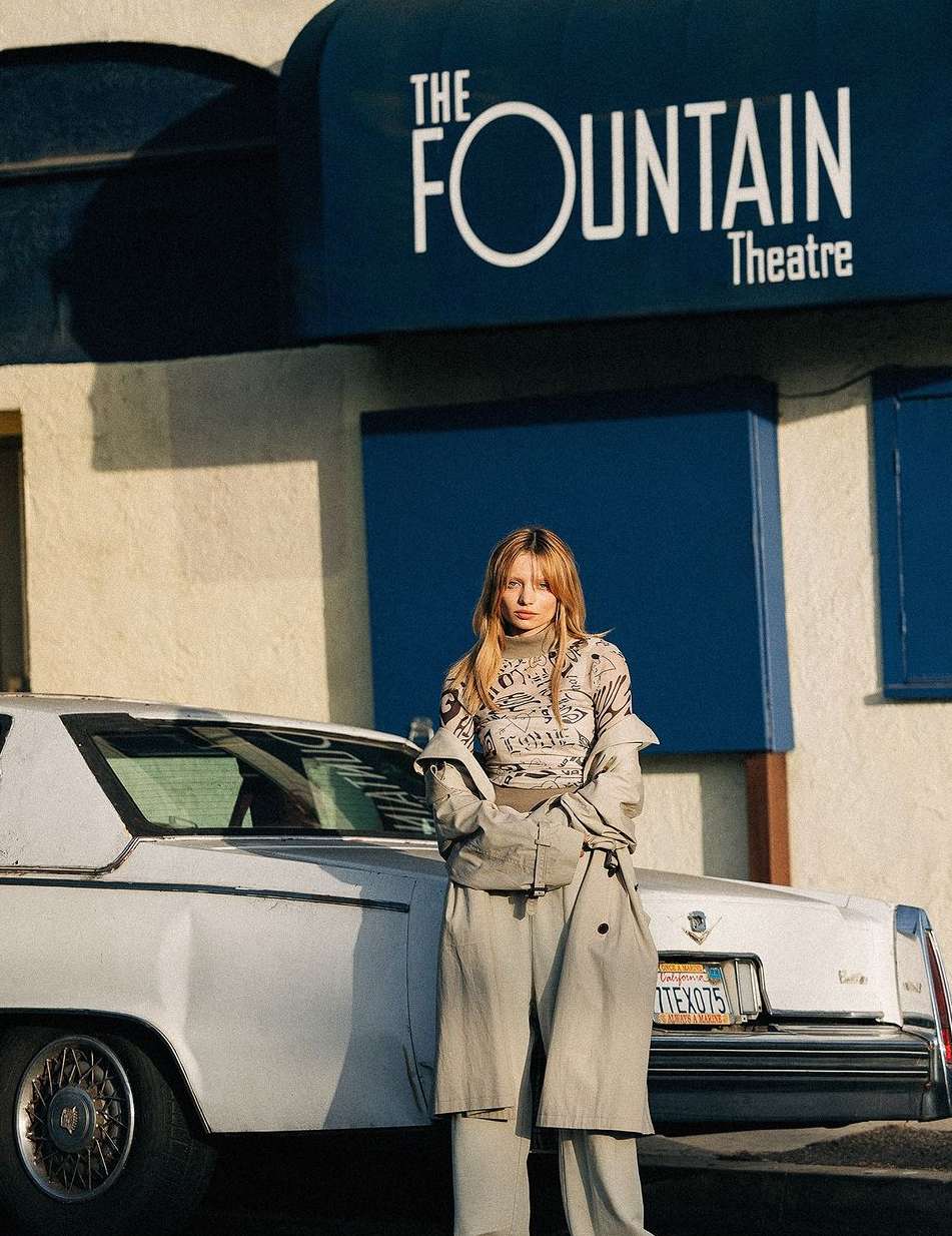 Adult female model in oversized trench coat and pants stands near a white car in front of The Fountain Theatre with blue awning.