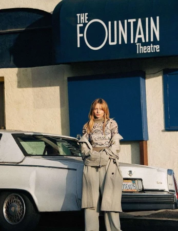 Adult female model in oversized trench coat and pants stands near a white car in front of The Fountain Theatre with blue awning.