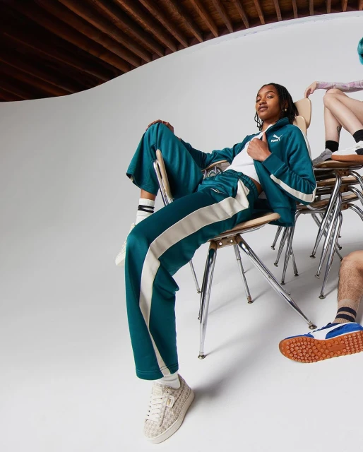 Two adult fashion models pose sitting on chairs in tracksuits, showcasing a modern apparel brand in a bright photo studio.