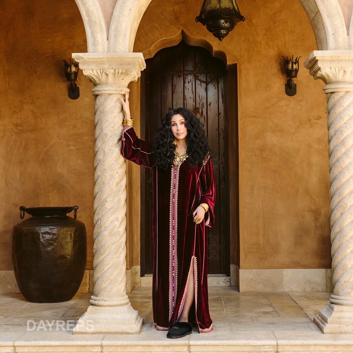 Cher in a maroon velvet dress with columns and an arched doorway with a lantern, exuding celebrity elegance and timeless style.