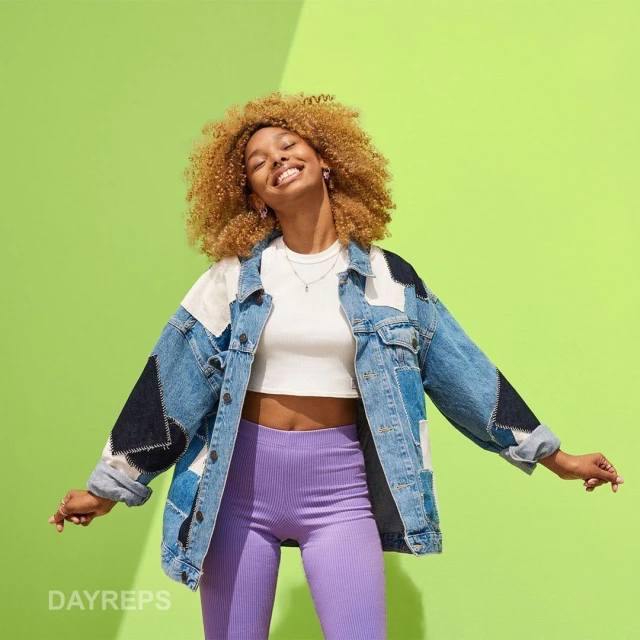 Smiling woman with curly hair wears a patchwork denim jacket and purple leggings against a bright green background, illuminated by studio lighting.