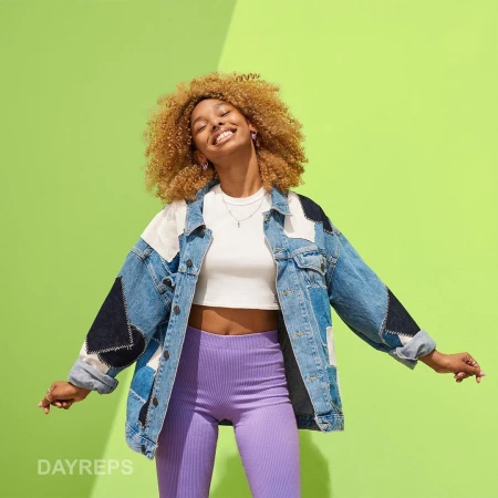Smiling woman with curly hair wears a patchwork denim jacket and purple leggings against a bright green background, illuminated by studio lighting.