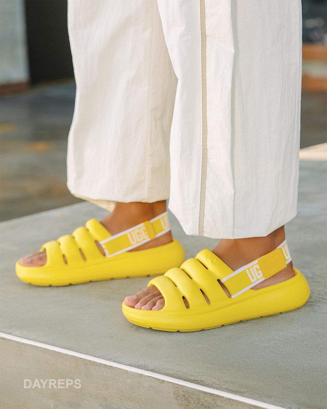 A close-up of yellow UGG sandals on a person wearing white pants, showcasing a casual fashion style and comfortable footwear.