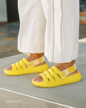 A close-up of yellow UGG sandals on a person wearing white pants, showcasing a casual fashion style and comfortable footwear.