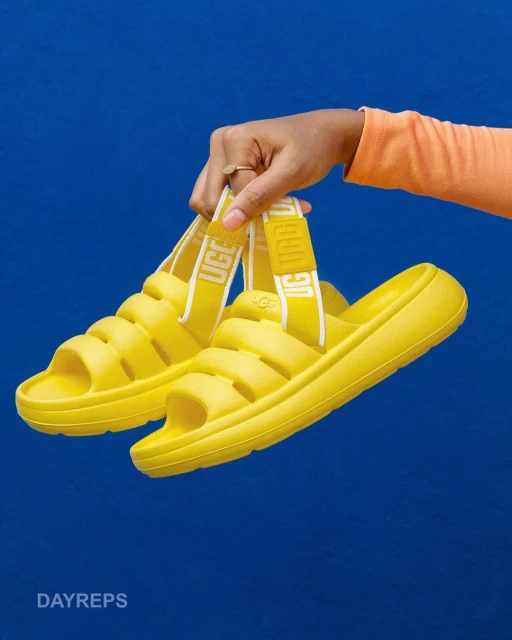 A pair of yellow UGG slides are held by a hand against a blue background, showcasing their unique design and vibrant color.