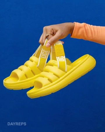A pair of yellow UGG slides are held by a hand against a blue background, showcasing their unique design and vibrant color.