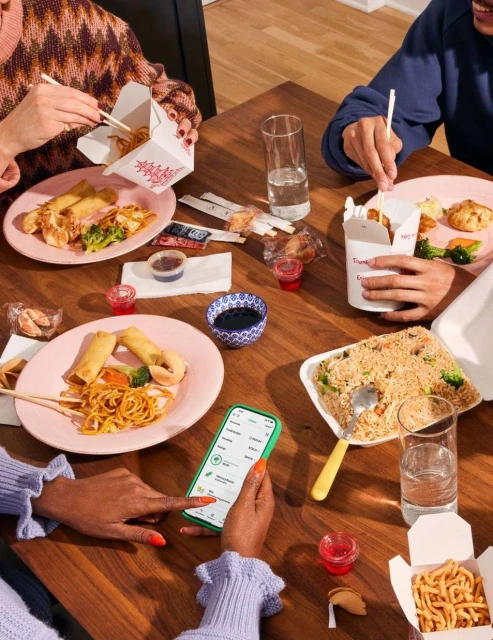 Overhead shot of four people at a wooden table enjoying a takeout meal with fried rice, noodles, egg rolls, and other dishes.