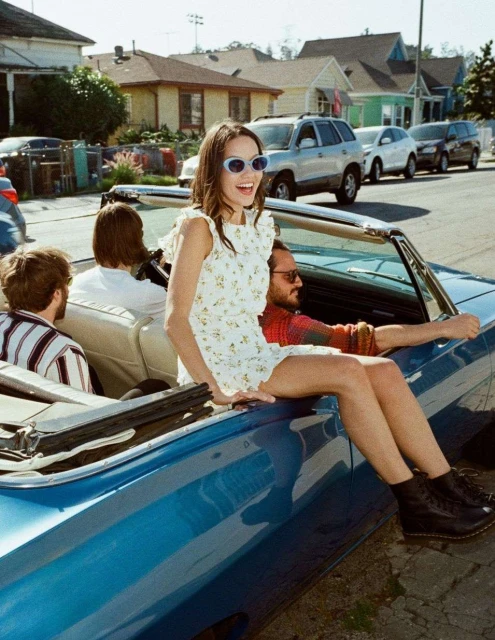 A smiling woman in a floral dress sits on a blue convertible in a residential neighborhood with three other people inside the car.