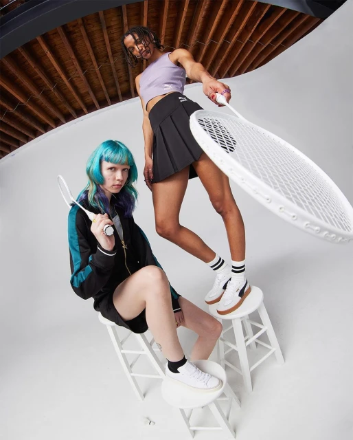 Two young adult women pose with tennis rackets in a bright studio. One sits on a white stool, the other stands, both in sports apparel.