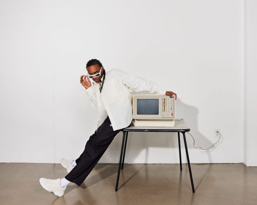 A man leans on a retro computer on a table in a minimalist setting, wearing a white shirt, dark pants, and white sunglasses.