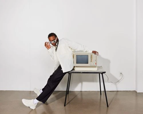 A man leans on a retro computer on a table in a minimalist setting, wearing a white shirt, dark pants, and white sunglasses.