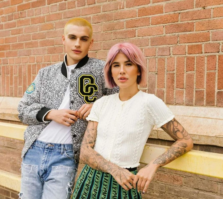 Two young adult models pose against a brick wall: one in a patterned jacket, the other in a white cardigan and patterned skirt.