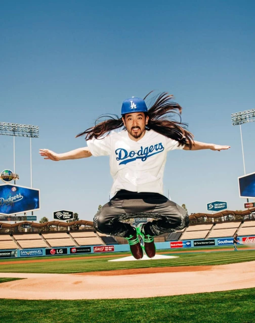 Man in Dodgers gear jumps high on a baseball field, arms outstretched, against a blue sky backdrop with stadium seating.