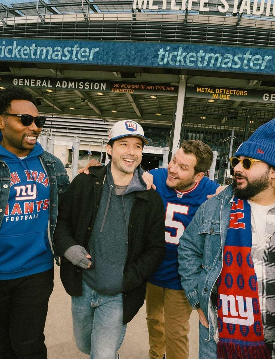 Four adult males are standing in front of MetLife Stadium, wearing New York Giants apparel. They are smiling and casually posing.