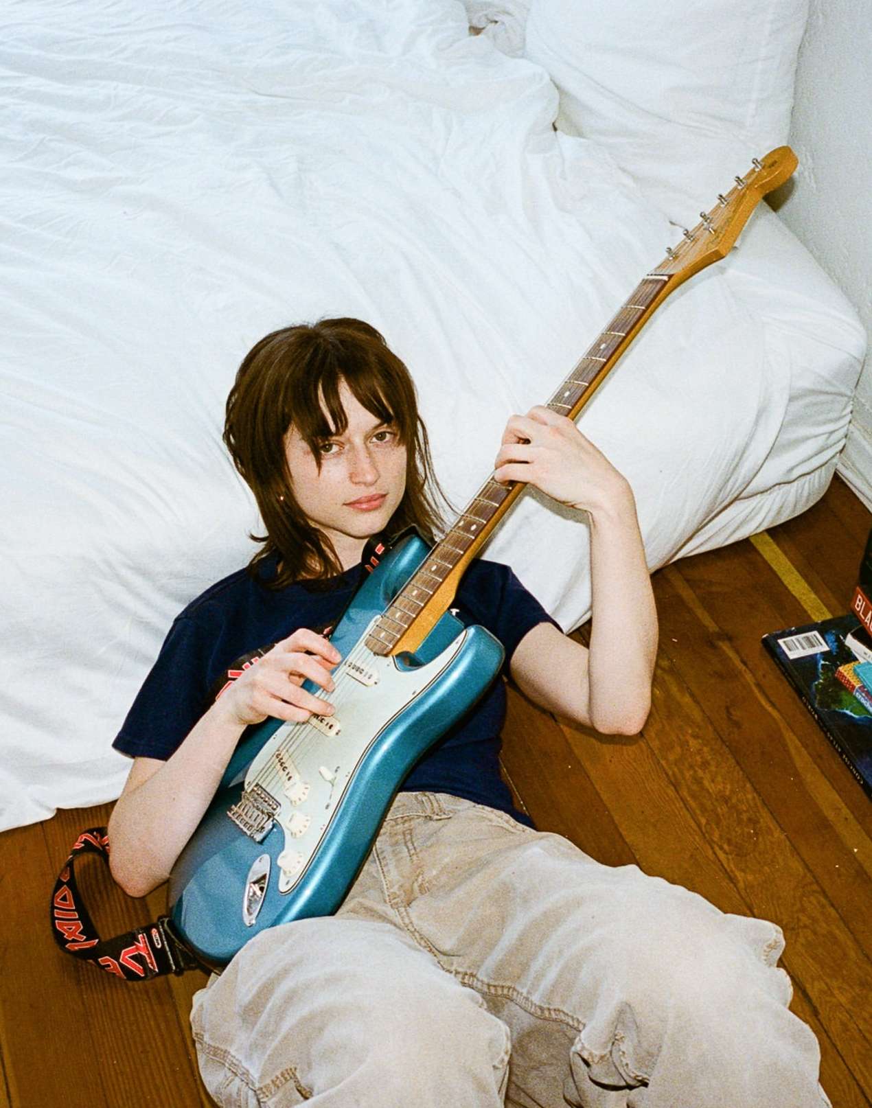 Young woman posing indoors holding an electric guitar, leaning against a white bed in casual clothing. Natural light illuminates the scene.